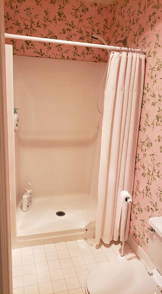 Small beige bathroom with a shower stall, white curtain, floral wallpaper, and white tile floor