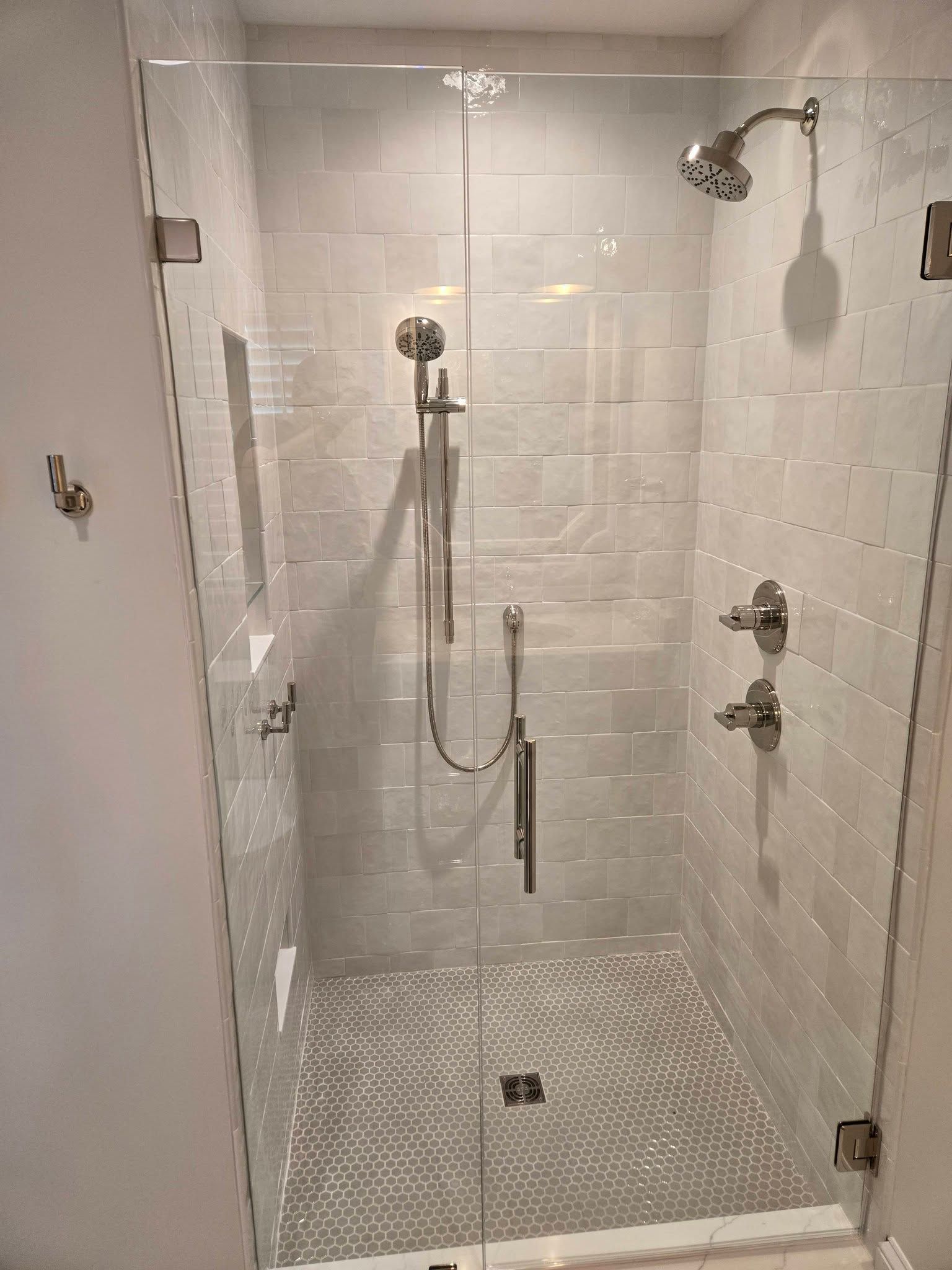 Small white tiled shower with handheld showerhead, grab bars, and a bench-style seat.