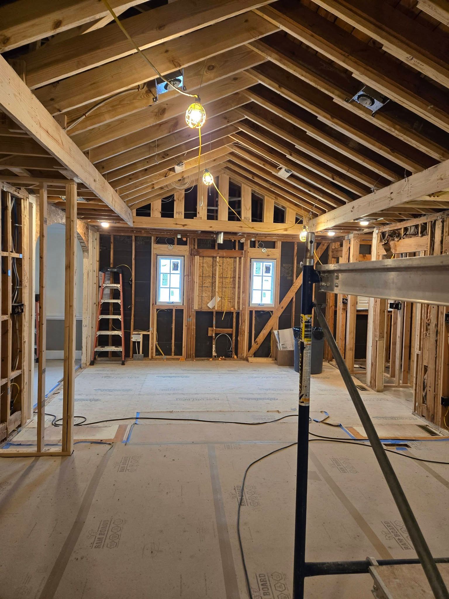 Unfinished attic renovation with exposed wooden rafters, insulation, tools, and ladder in a narrow room.
