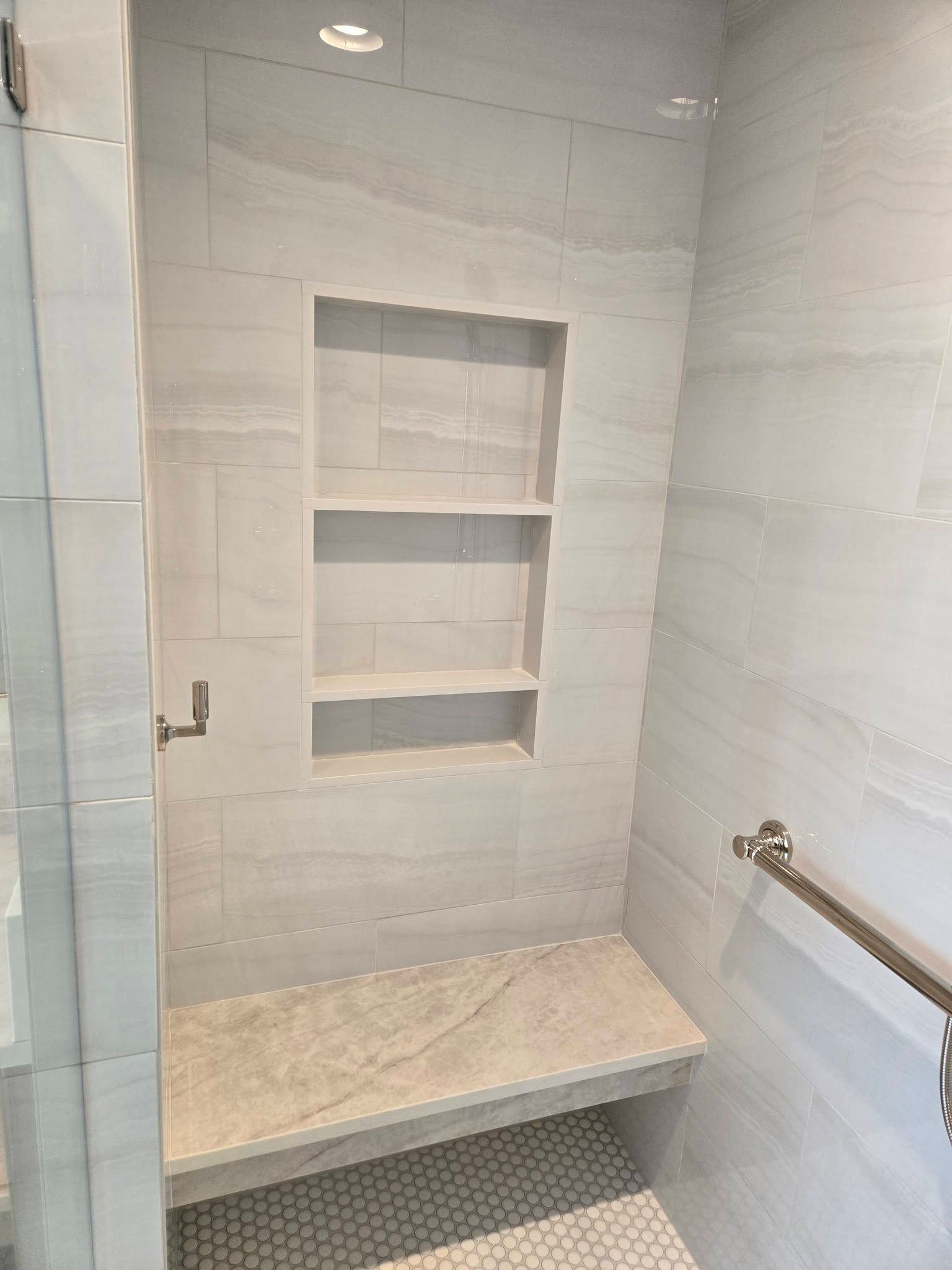 Shower alcove with built-in shelves, stone bench, and grab bar in a tiled bathroom