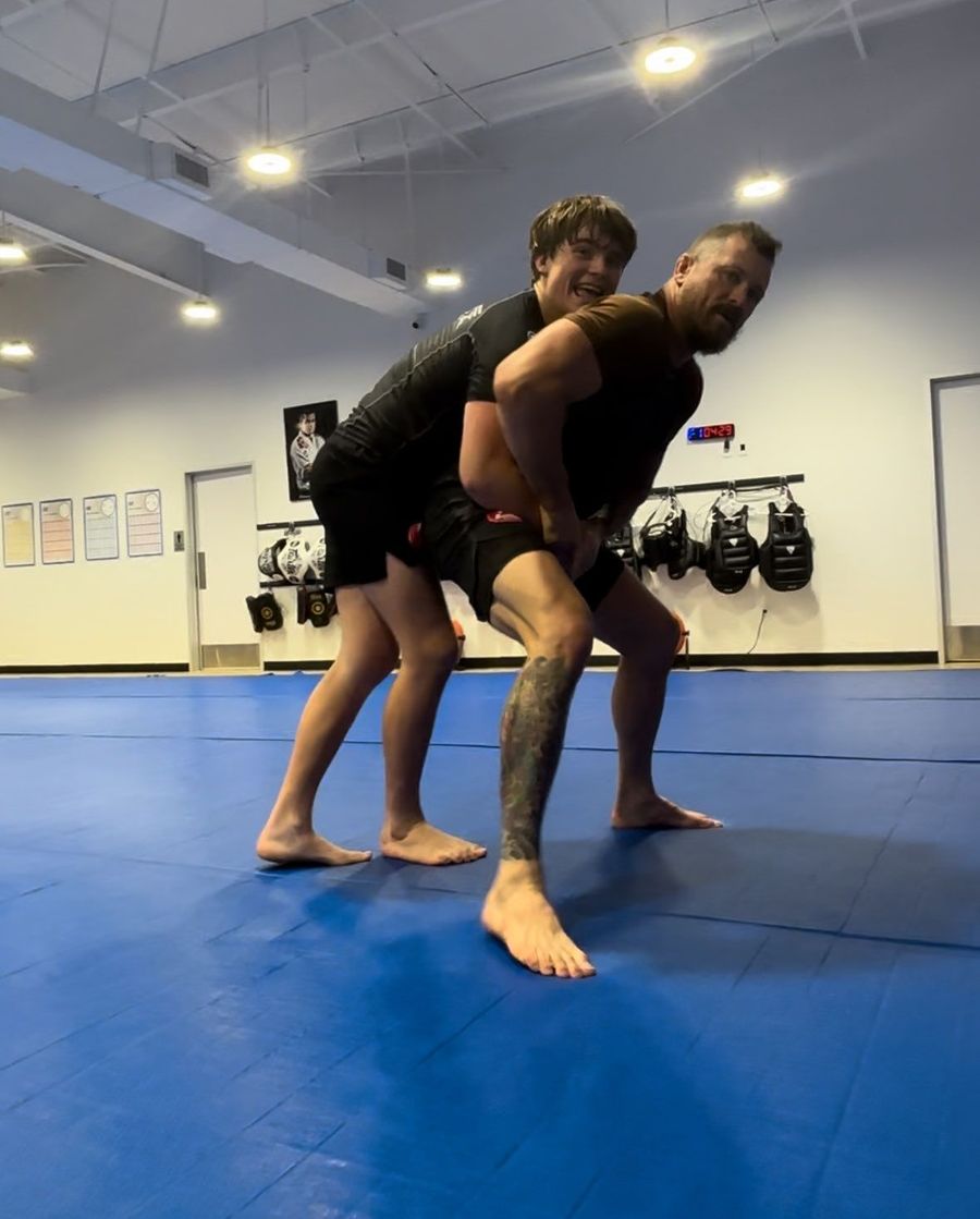 Two people grappling on blue mats in Gracie Barra Sienna dojo in Fresno, TX, with wall-mounted gear in the background.