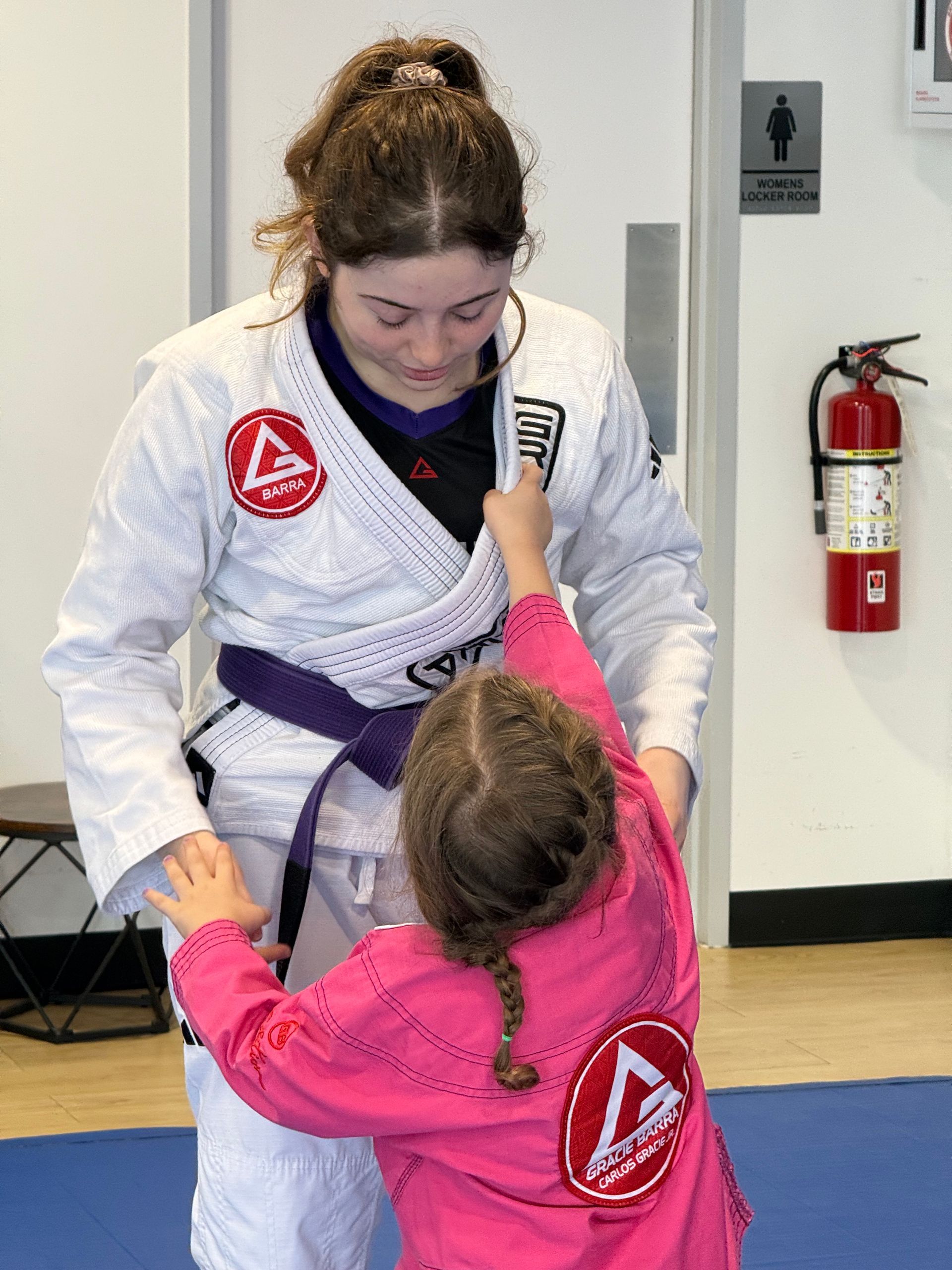 Two judo practitioners in gis, one helping the other adjust a belt in Gracie Barra Sienna dojo in Fresno, TX