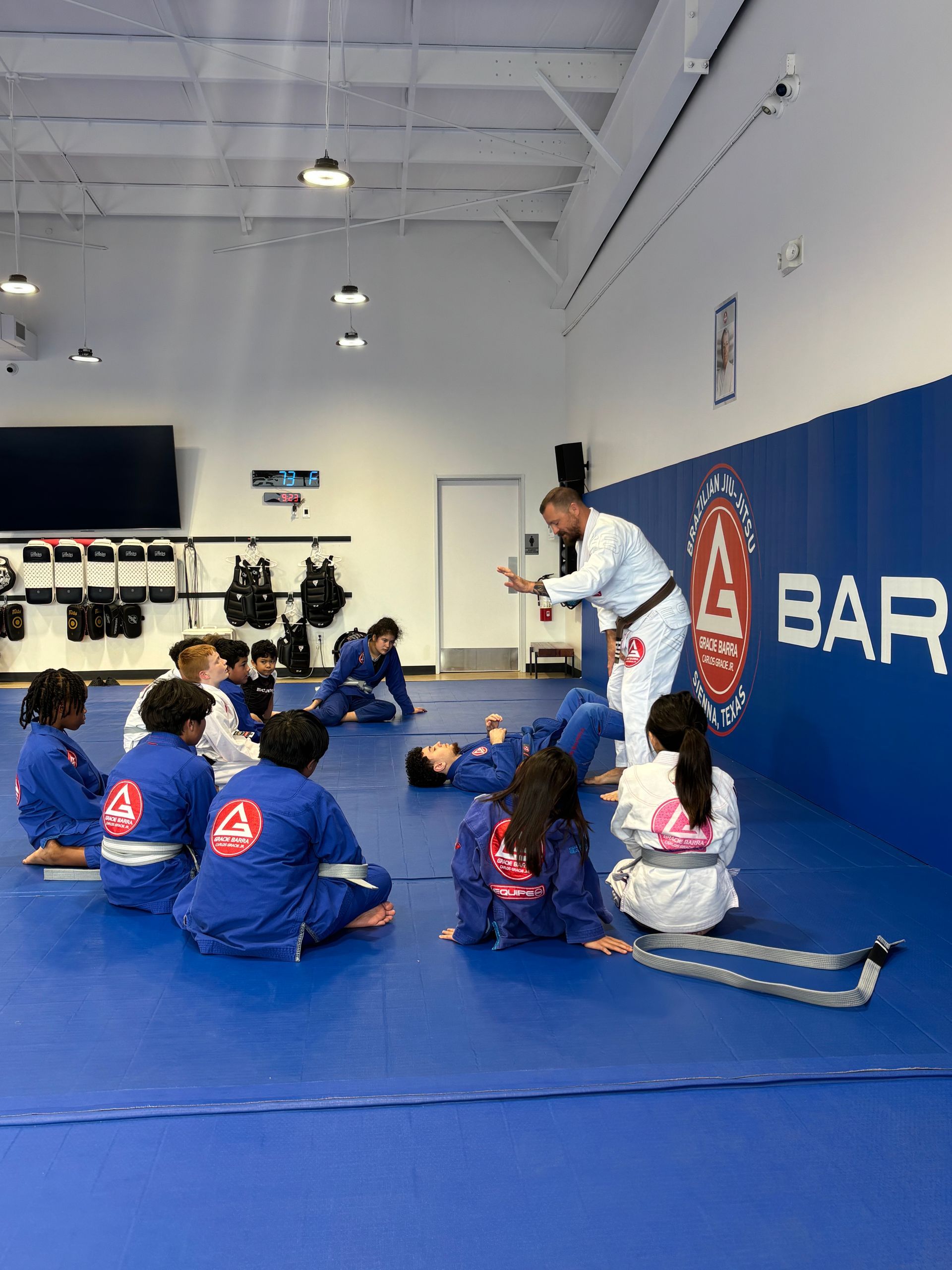Students in blue gis sit on a mat as an instructor speaks in Gracie Barra Sienna dojo in Fresno, TX