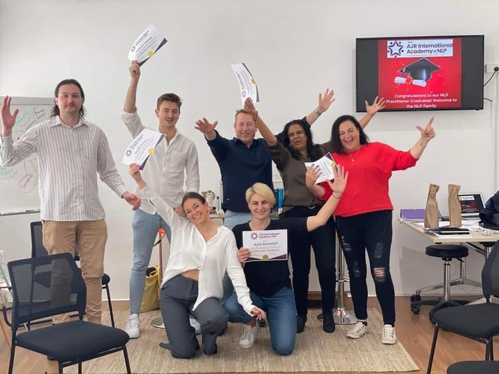Group of people smiling and waving in an office, holding flyers and certificates near a wall-mounted screen.