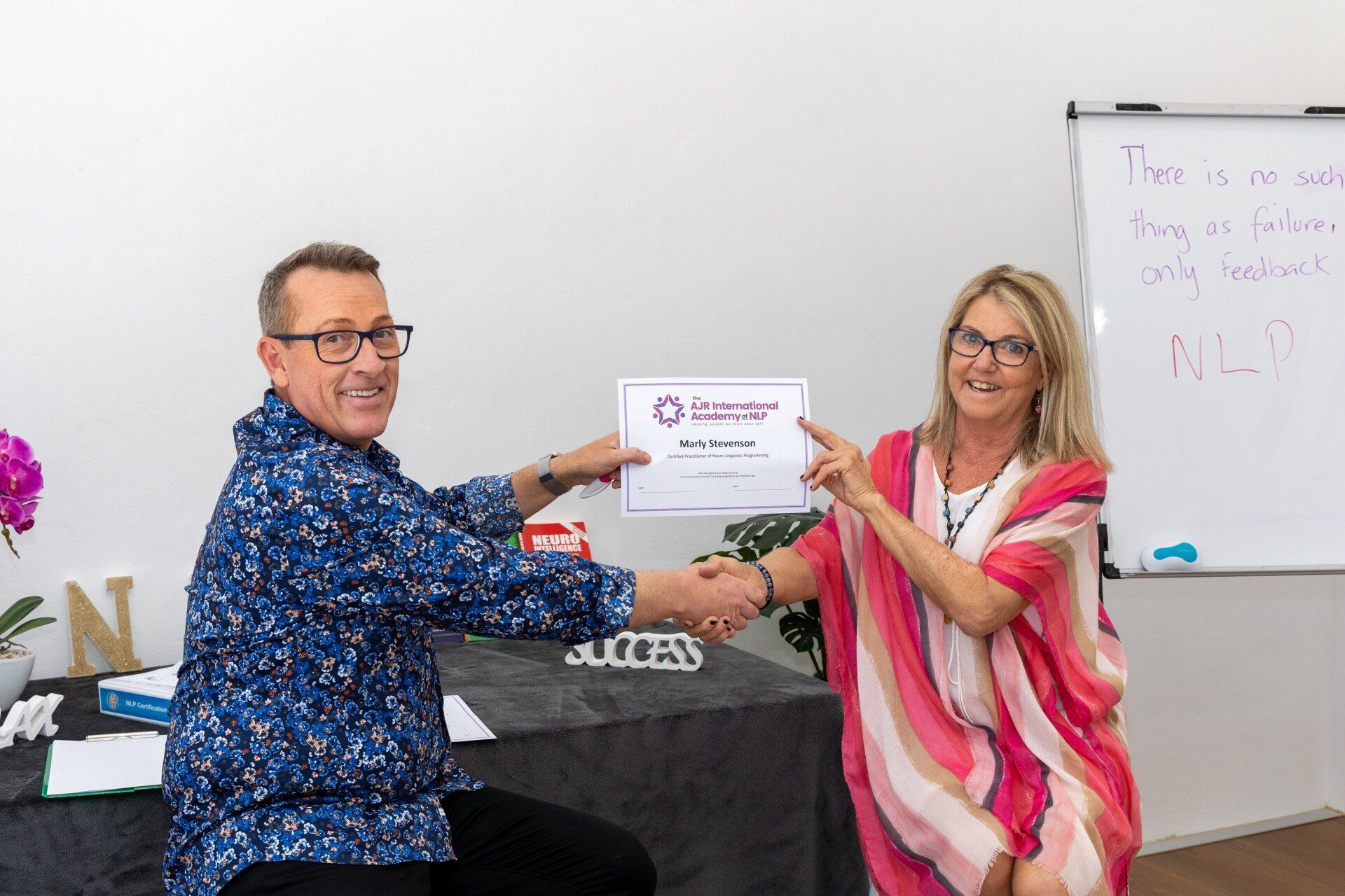 Two people smiling and holding a certificate in a bright room with a whiteboard.