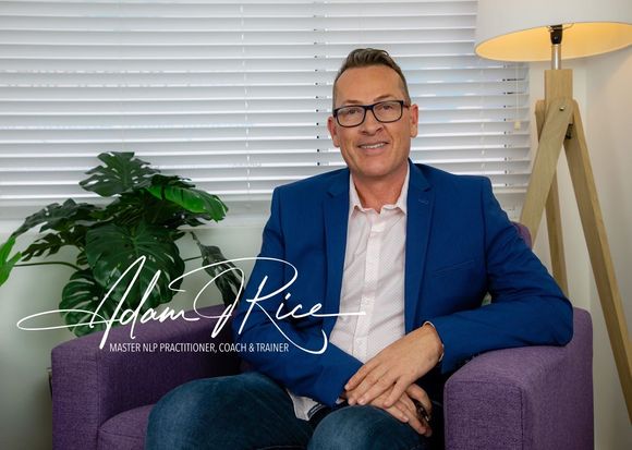 Man sitting on a purple couch in a bright office, smiling beside a plant and floor lamp.