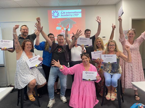 Group of smiling people on a stage, holding certificates and raising hands in a colorful classroom event.