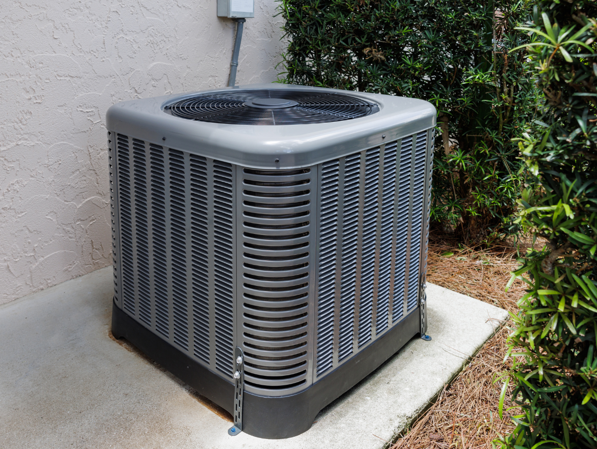 Air conditioning unit outside a building, gray and rectangular, near green shrubs.