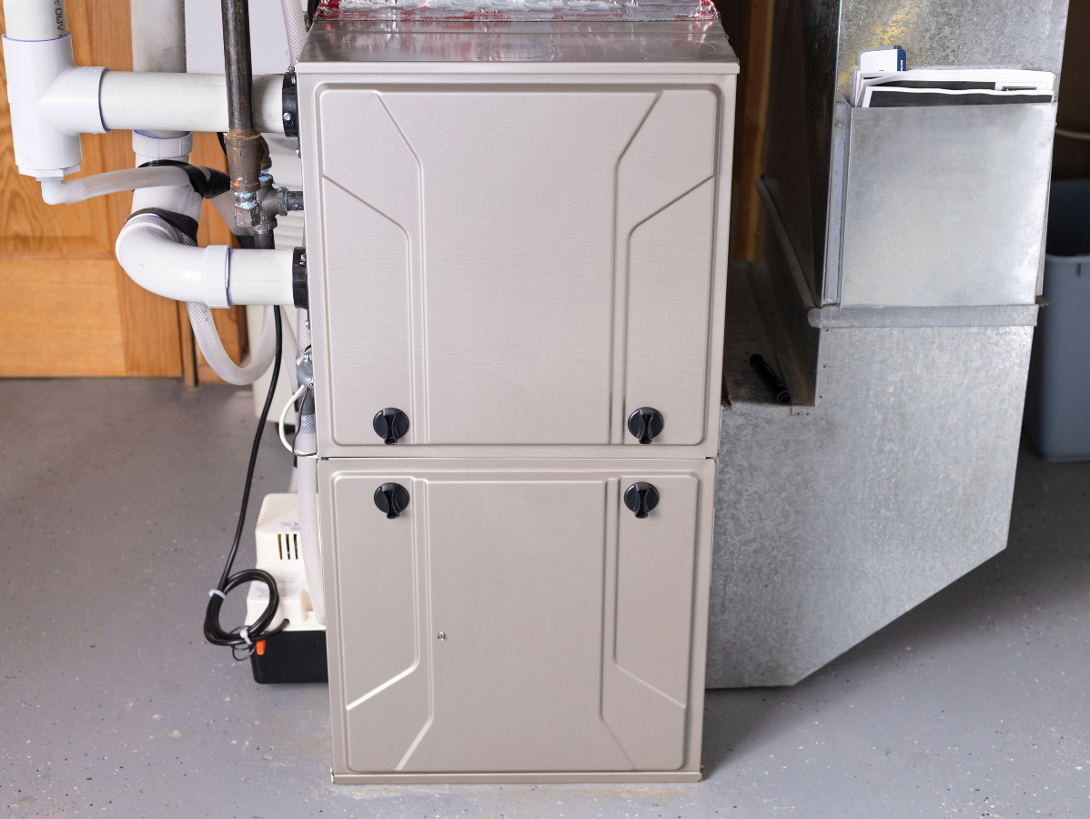 Tan furnace with two doors, surrounded by ducts and pipes in a utility room.