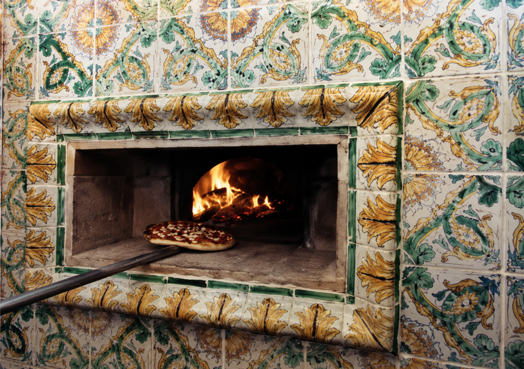Forno a legna incassato in una parete piastrellata, con le fiamme all'interno e una pizza che cuoce sulla pietra.