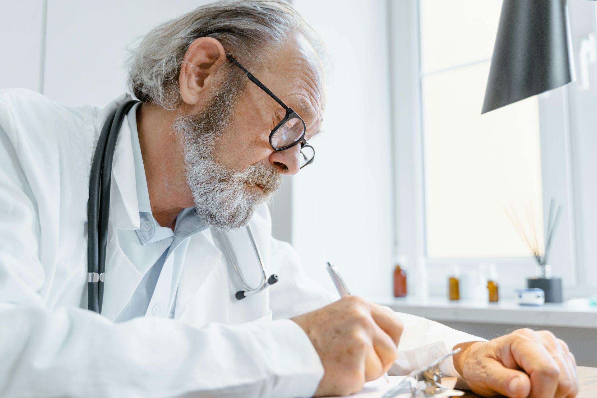 An older doctor is sitting at a desk writing on a clipboard.