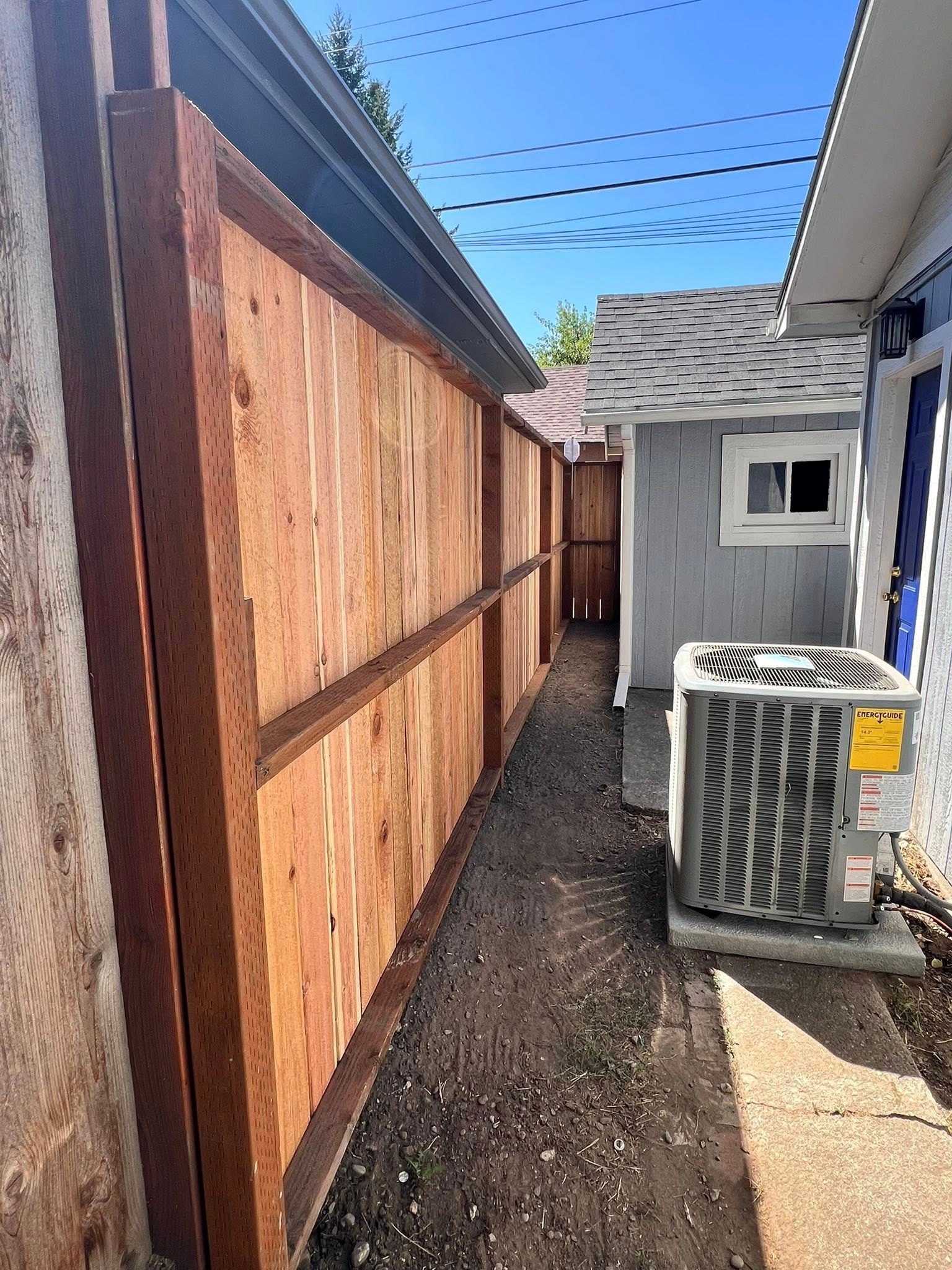 Wooden fence next to a building, a narrow gravel path in between. An AC unit is visible.