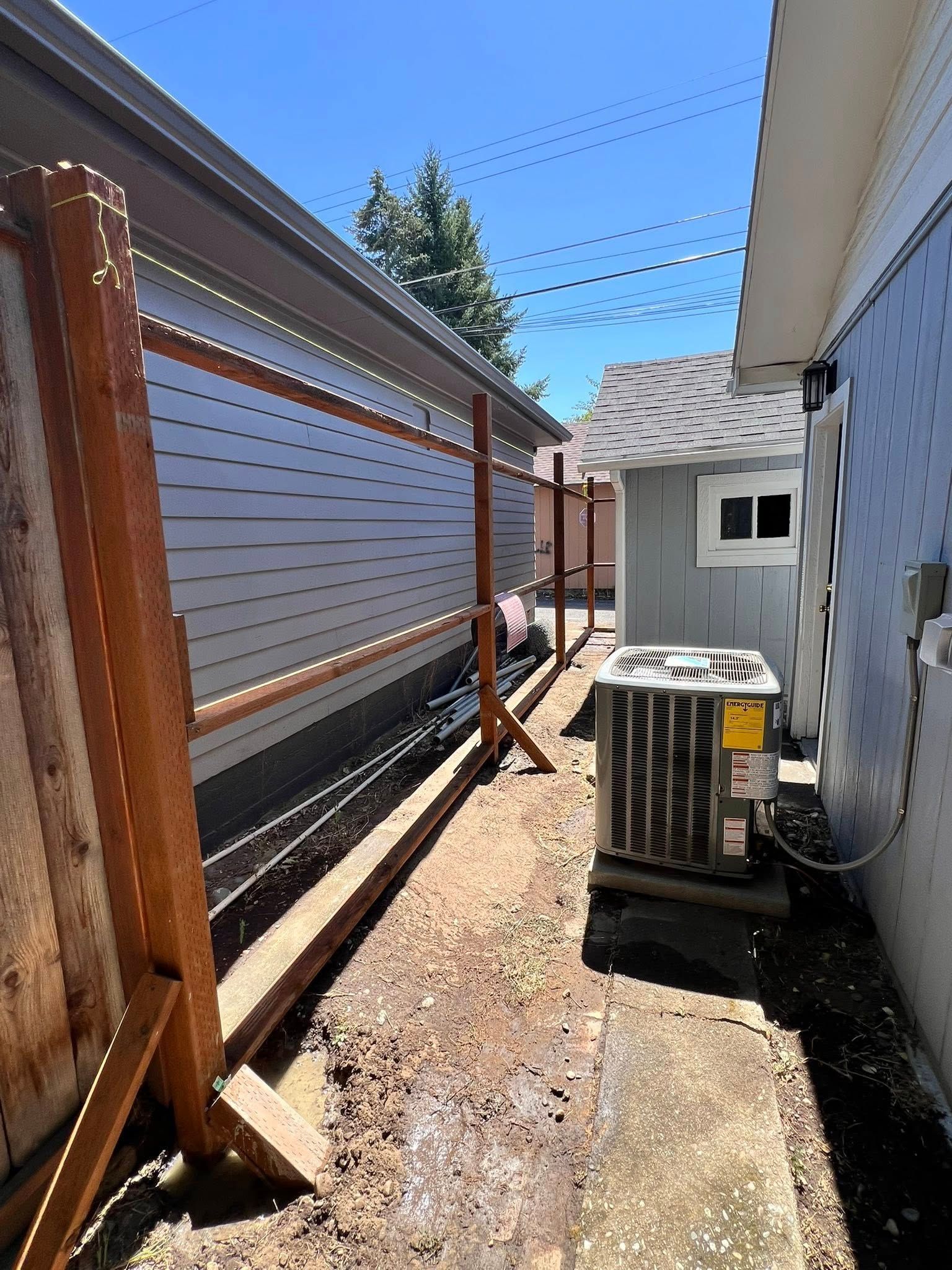 Narrow pathway between buildings, one with an AC unit. Wooden fence on the left, blue building on the right, clear sky.