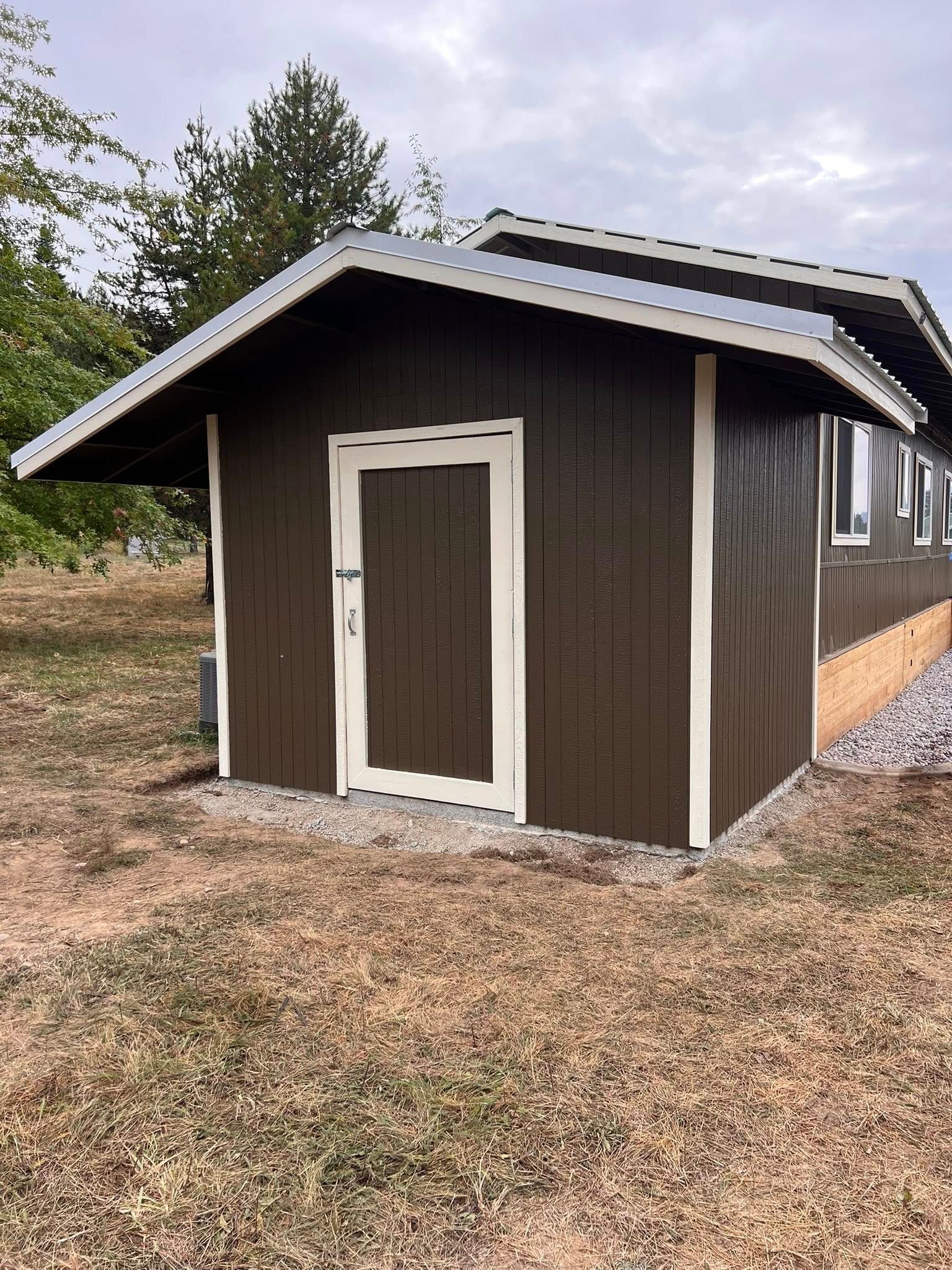 Dark brown shed with white trim, door, and metal roof edge sits on gravel and dry grass.