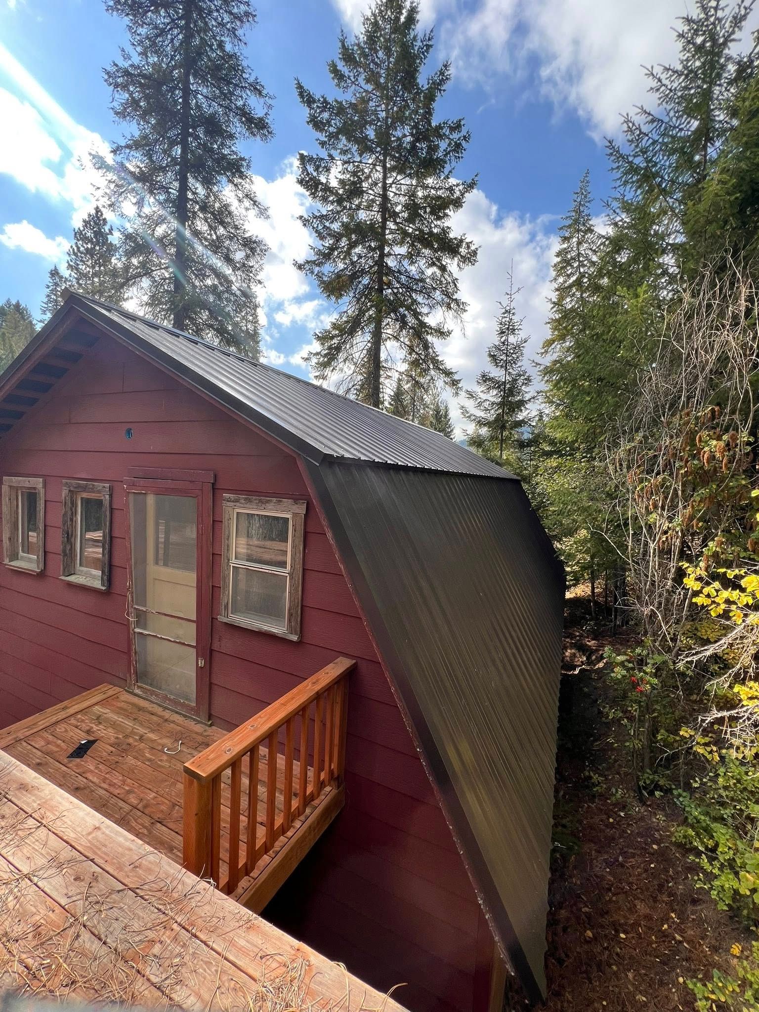 Red cabin with wooden deck, black roof, and trees in background under a cloudy blue sky.