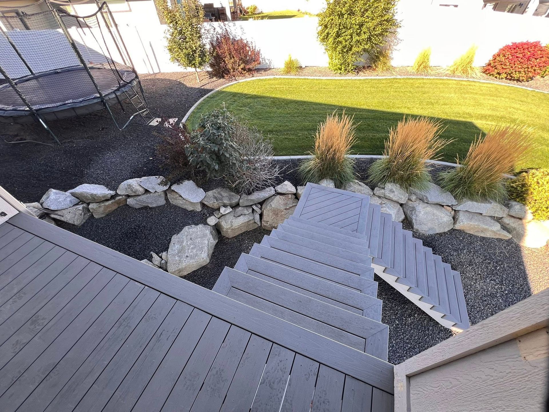 Gray deck steps lead down to a landscaped backyard with a trampoline and rock border.