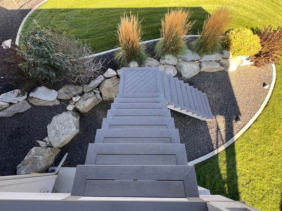 Gray staircase descends from a patio into a landscaped area with grass, rocks, and ornamental plants.
