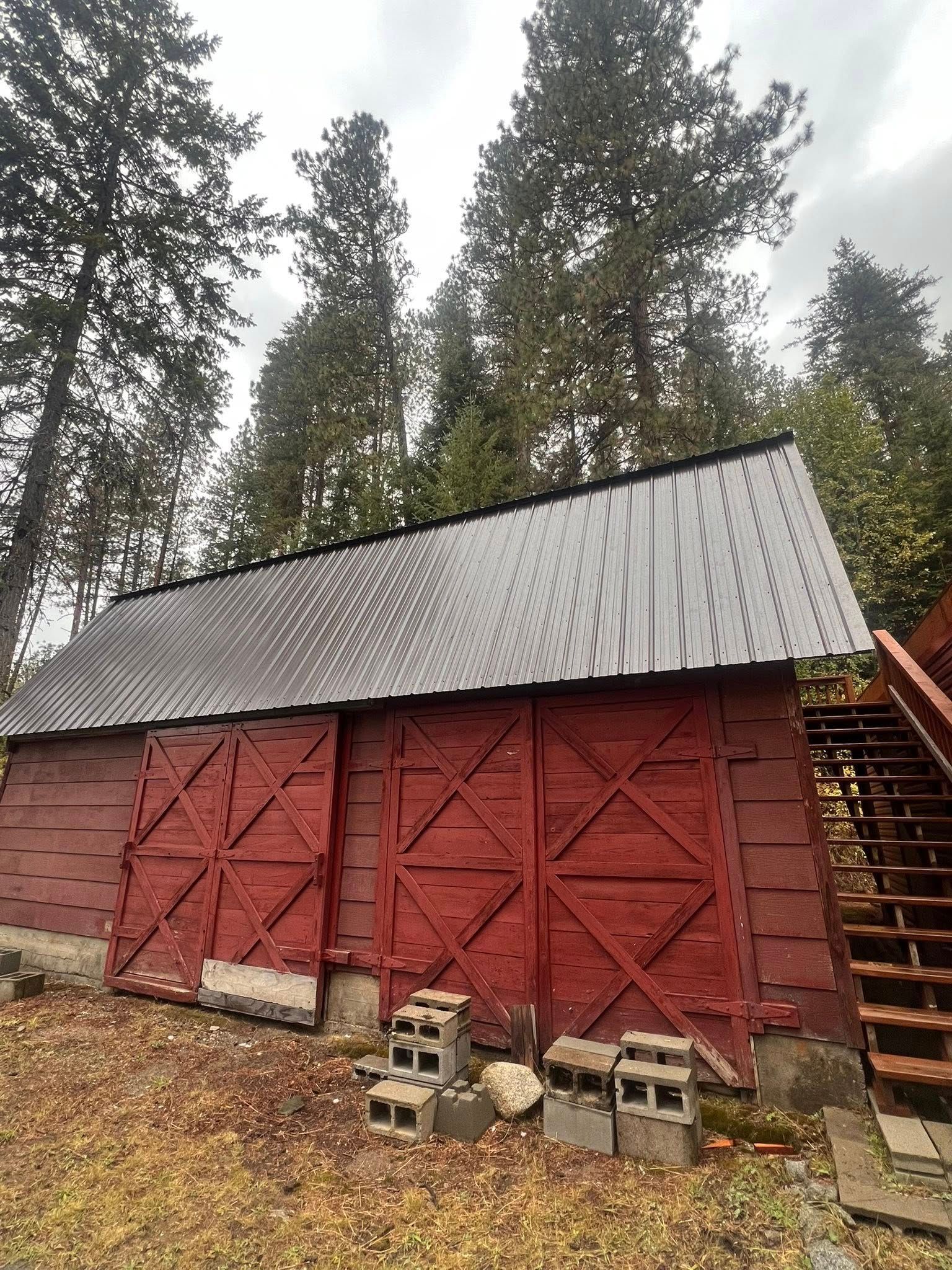 Red barn with a metal roof, tall trees in the background. Several cinder blocks are in front of the building.