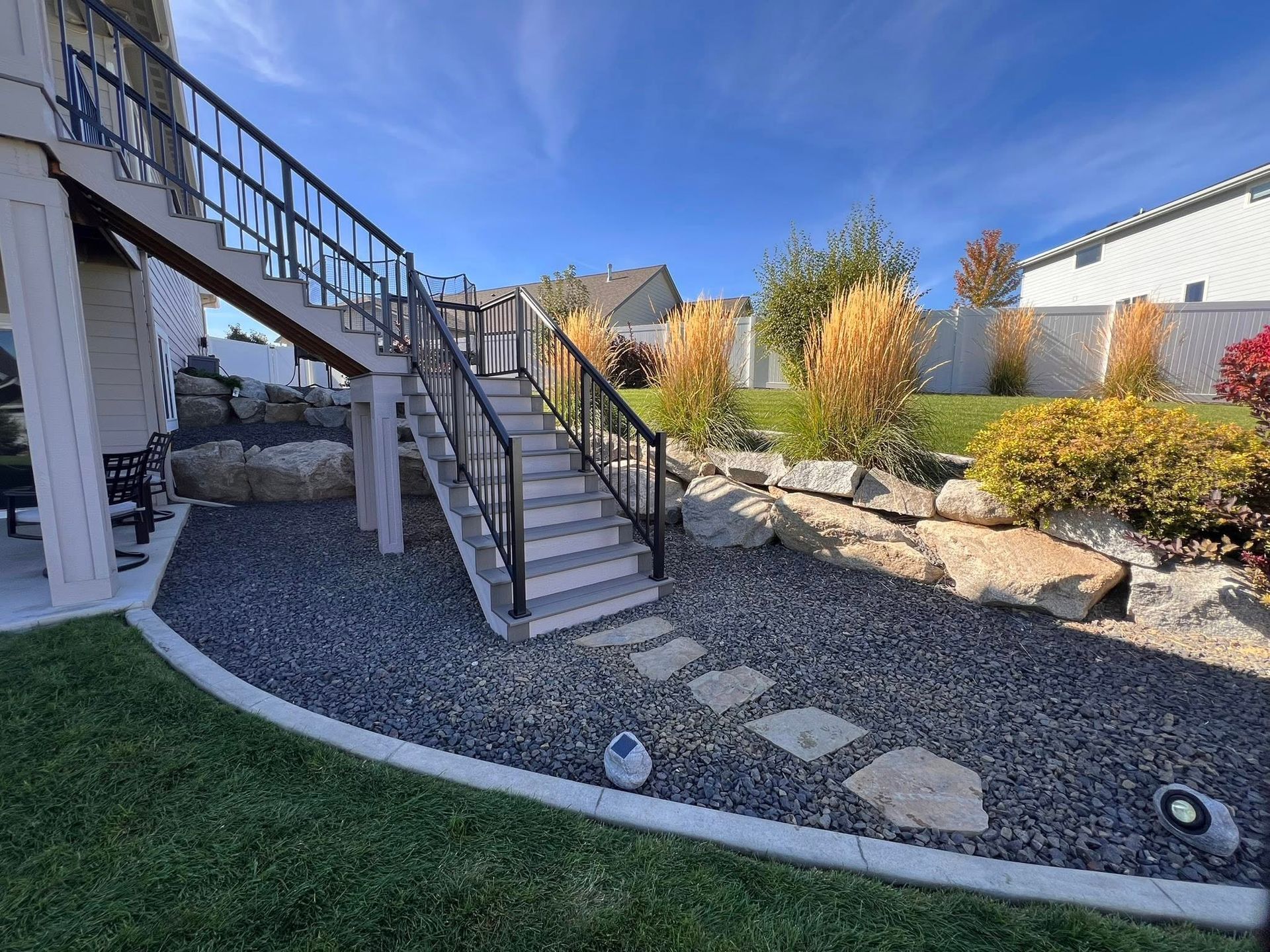 Stairs leading up to a deck with a black railing, set in a backyard with landscaping and a blue sky.