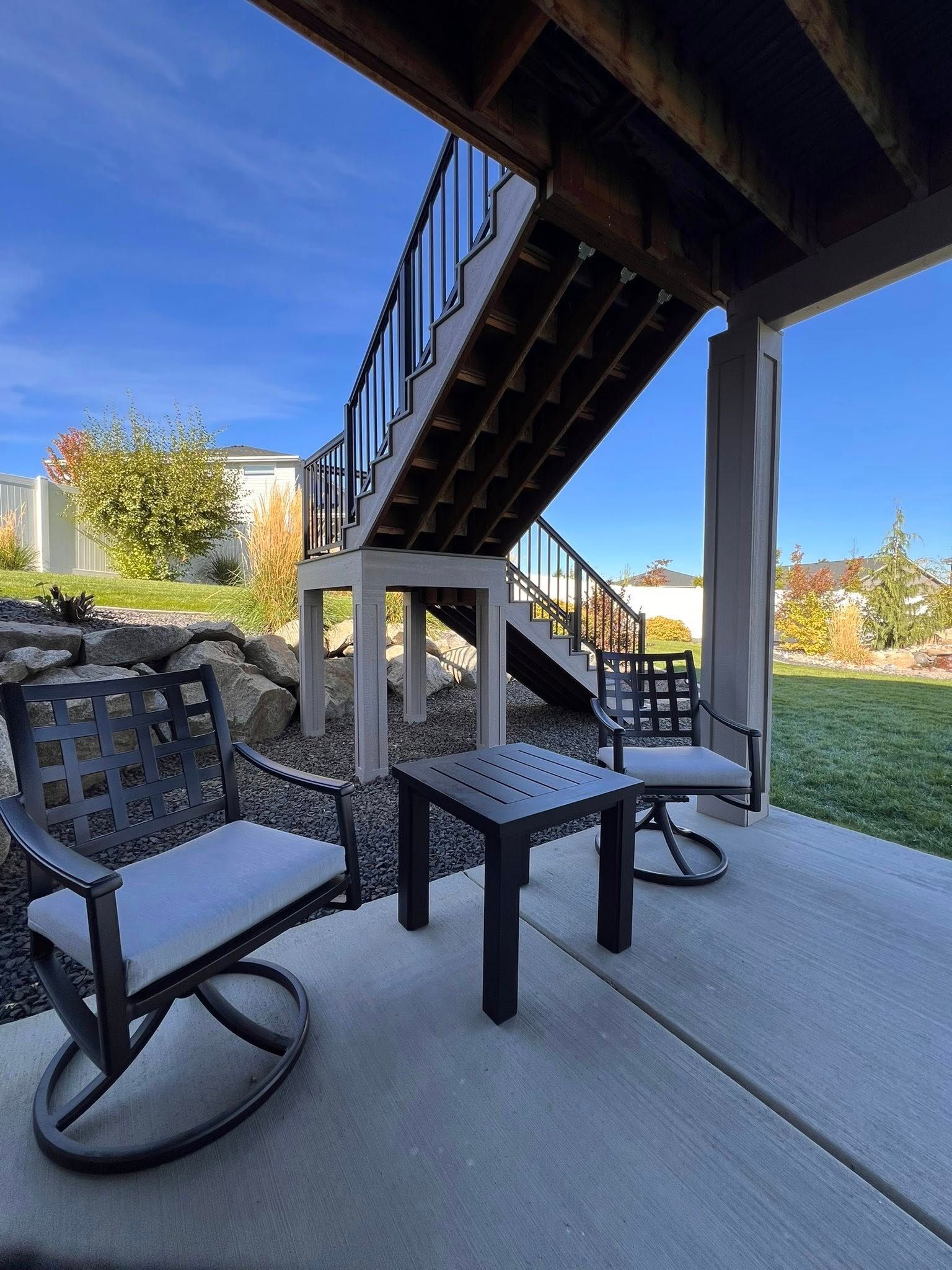 Patio with two chairs, small table under deck stairs. Concrete, grass, and a bright blue sky.