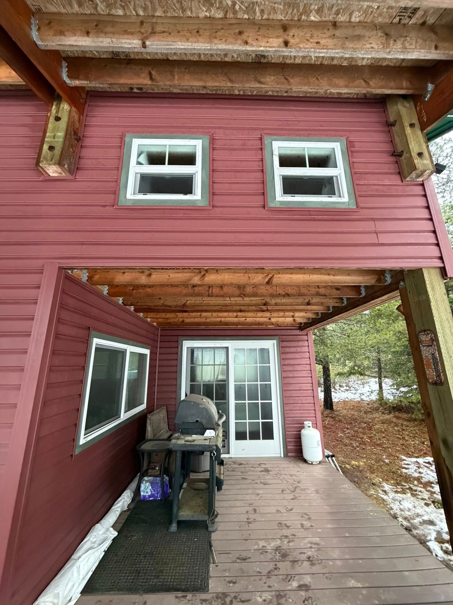 Red cabin with two windows, a sliding door, and a covered porch. A grill and propane tank are visible.