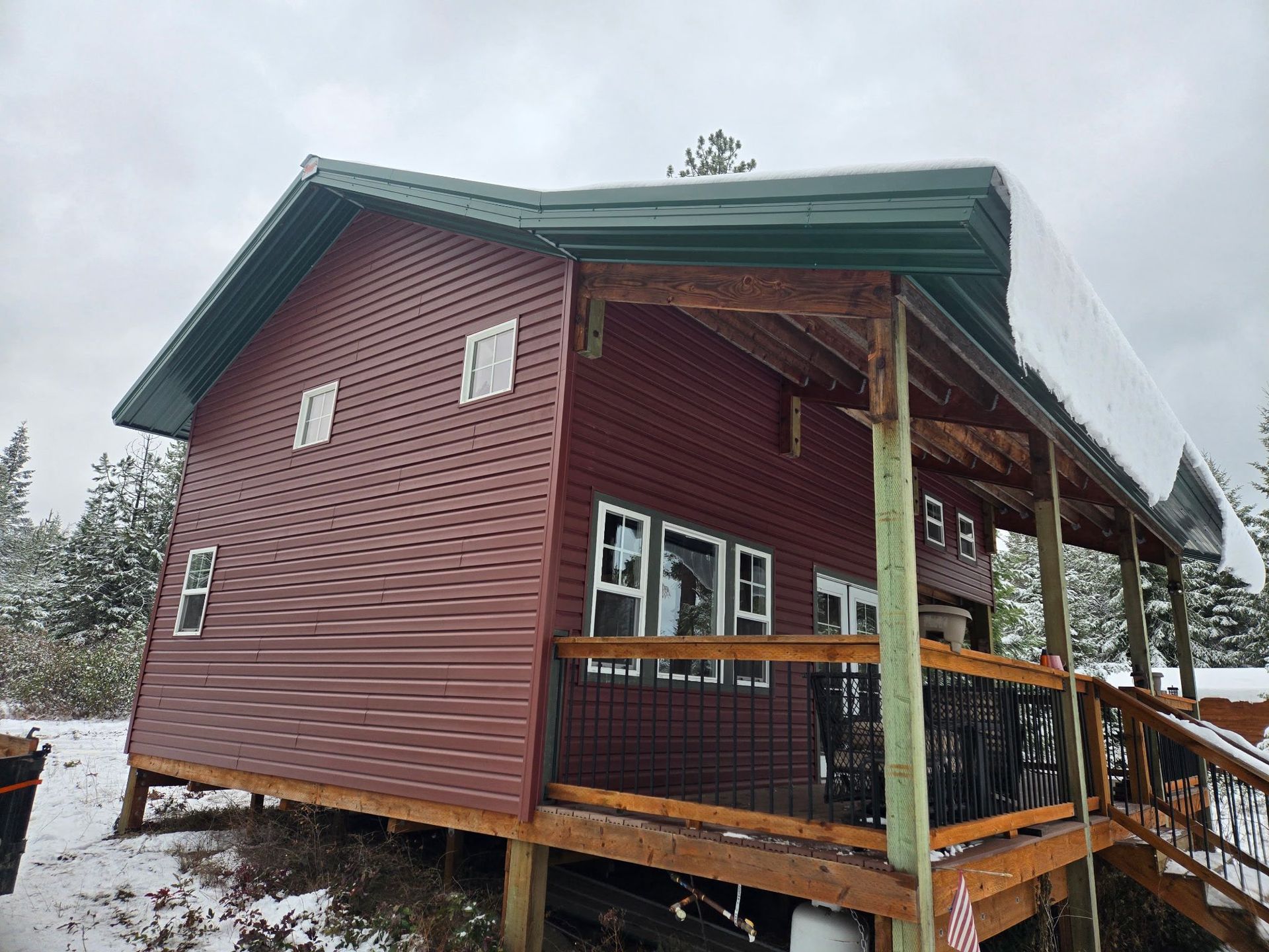 A maroon cabin with a green roof and a covered porch, set in a snowy outdoor environment.