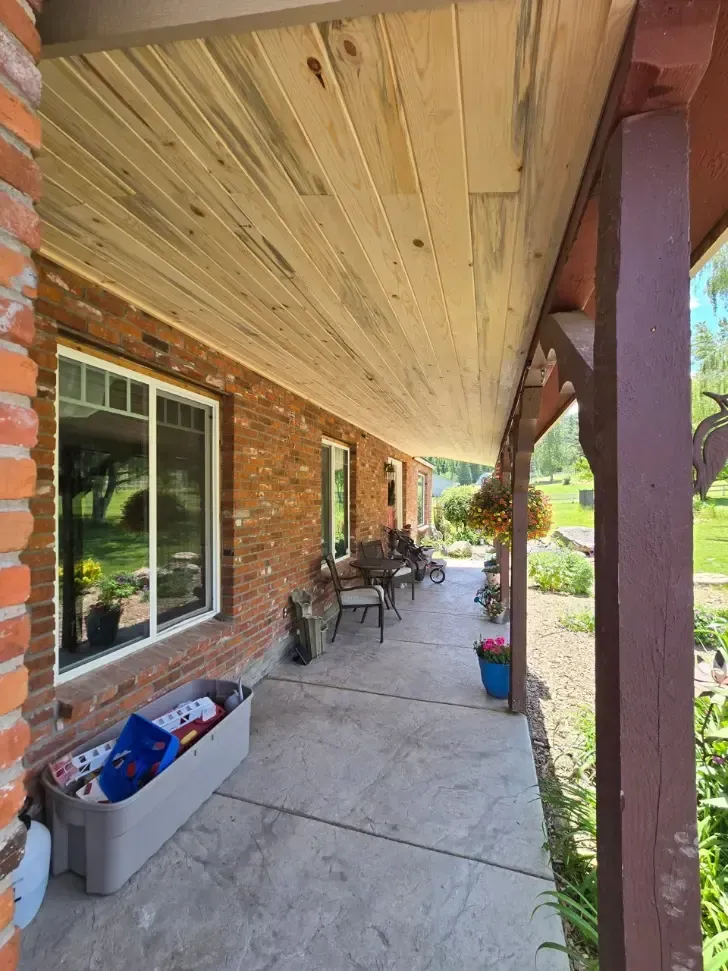 Covered porch with brick wall, concrete floor, and wooden ceiling. Furniture and plants line the porch.