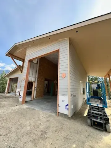 Exterior view of a house under construction with a lift. Light gray siding, tan wood trim, and a blue sky.
