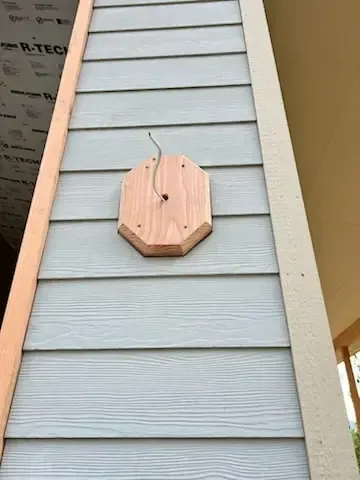 Wooden mounting block attached to gray siding, with exposed wire. Building exterior.