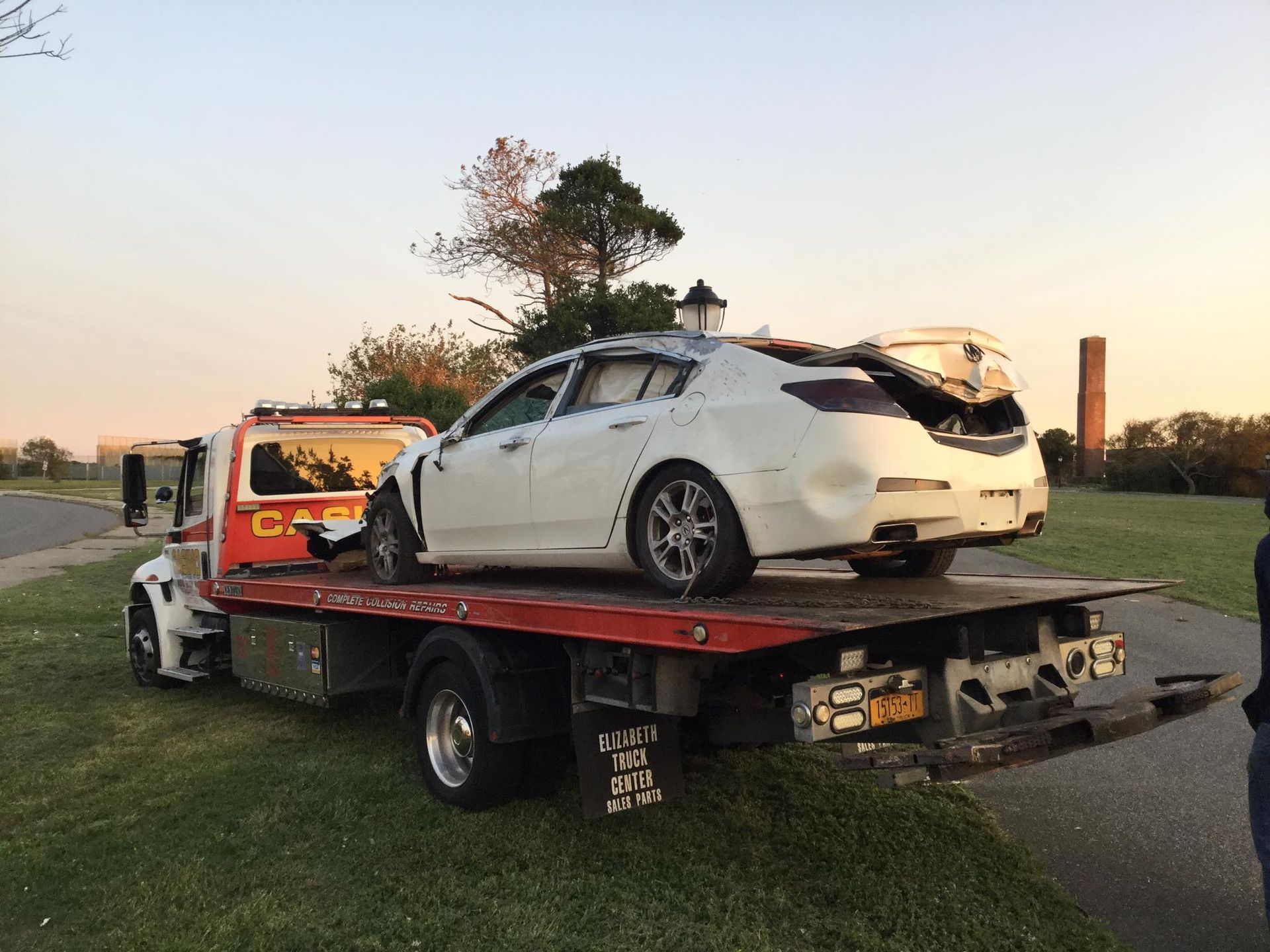 White sports car with rear damage loaded on a tow truck at sunset by a roadside park