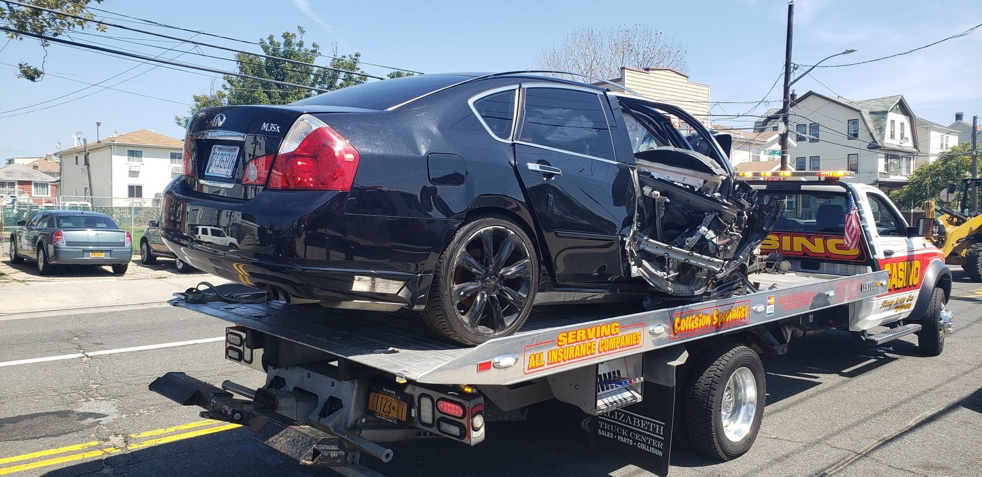 Black sedan on a tow truck with heavy front-end damage on a street.