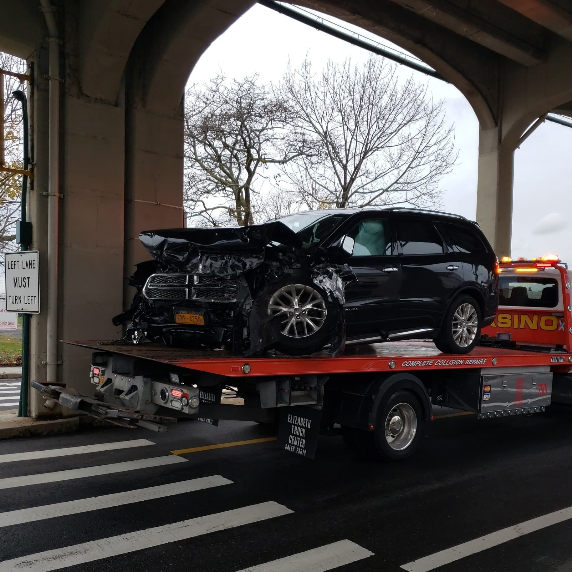Severely damaged black SUV on a tow truck under a bridge, with front end crushed.