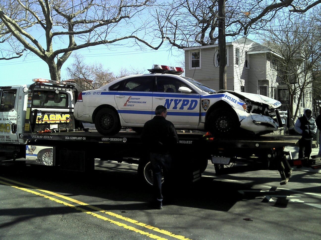NYPD police car loaded on a tow truck on a tree-lined street, with a person standing nearby