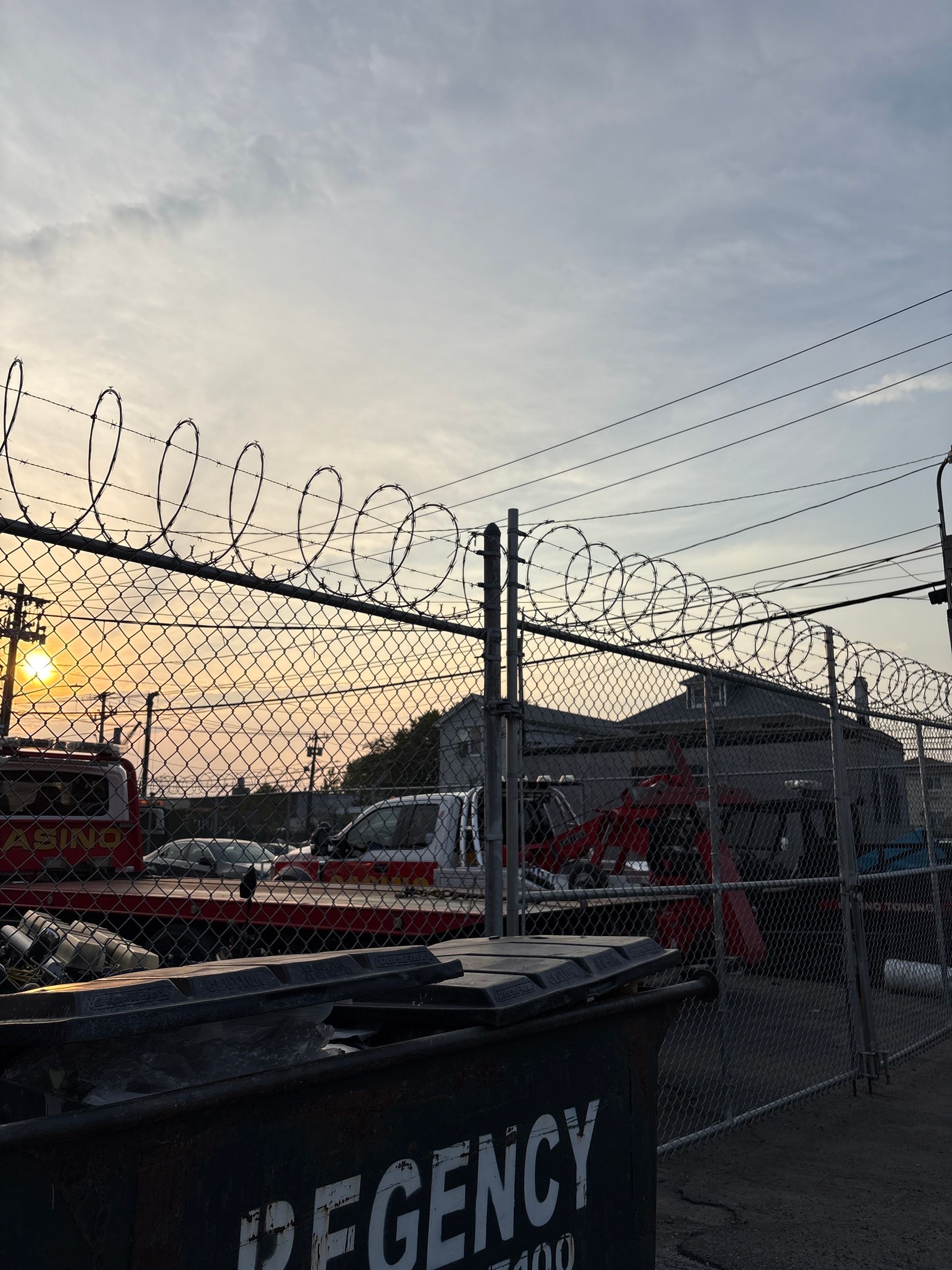 Barbed-wire fence at sunset with parked vehicles and a black dumpster labeled “REGENCY” in front
