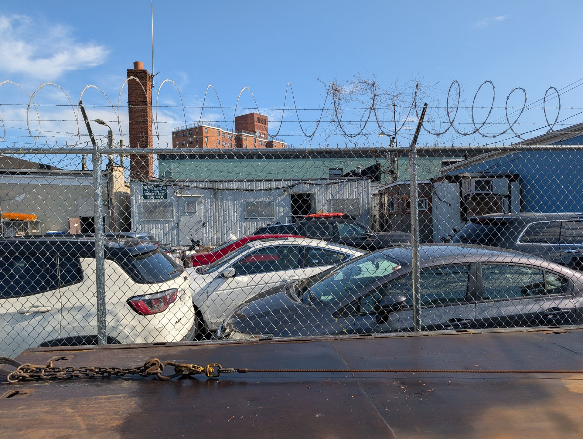 Chain-link fence topped with barbed wire in front of parked cars and industrial buildings.