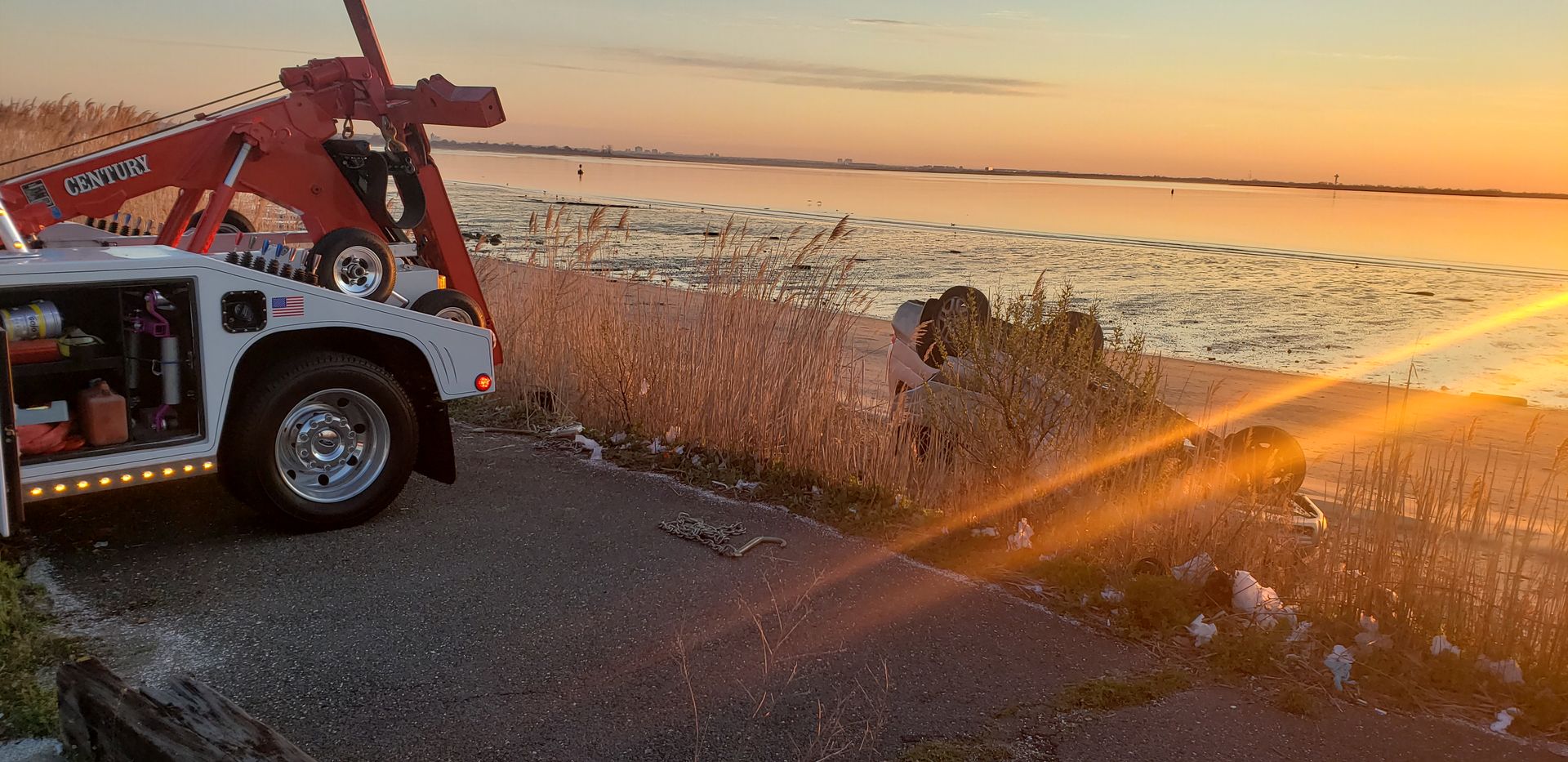 Tow truck by a lakeshore at sunset, with a dramatic orange sky and water beyond the road