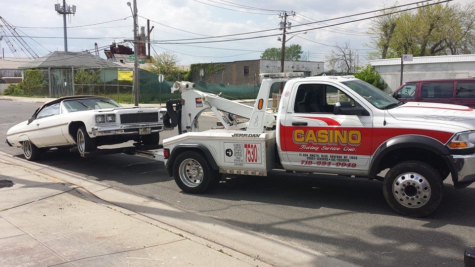 Tow truck towing a damaged white car on a roadside near power lines and houses