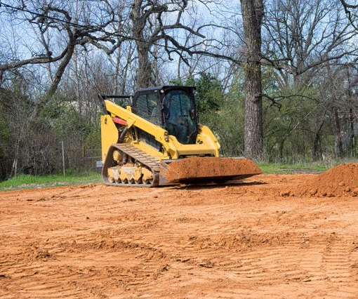 A person operating a small excavator, digging a trench in a wooded area with brown soil.