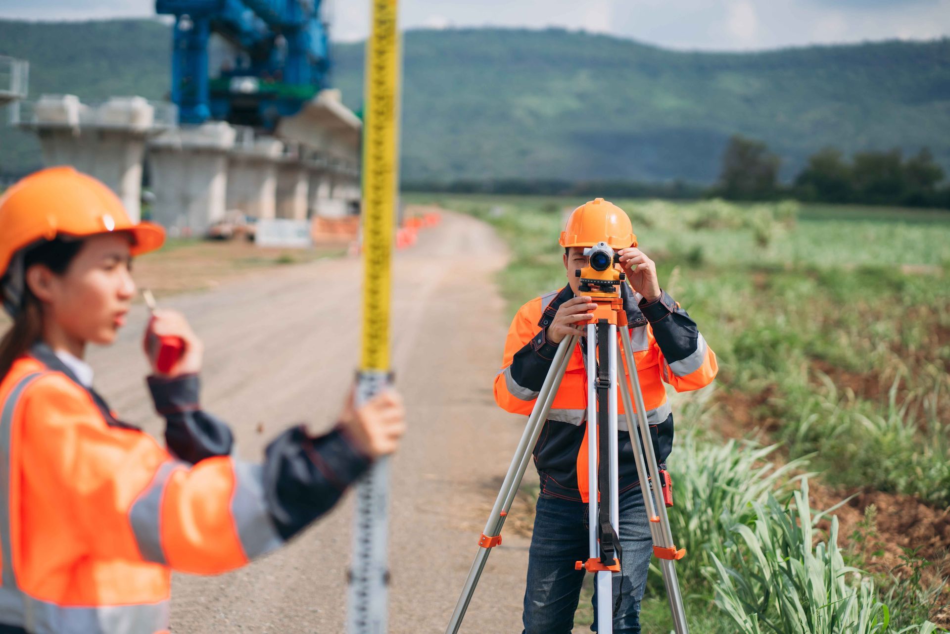 Surveyors in orange safety gear using tripods and a measuring rod along a rural roadside