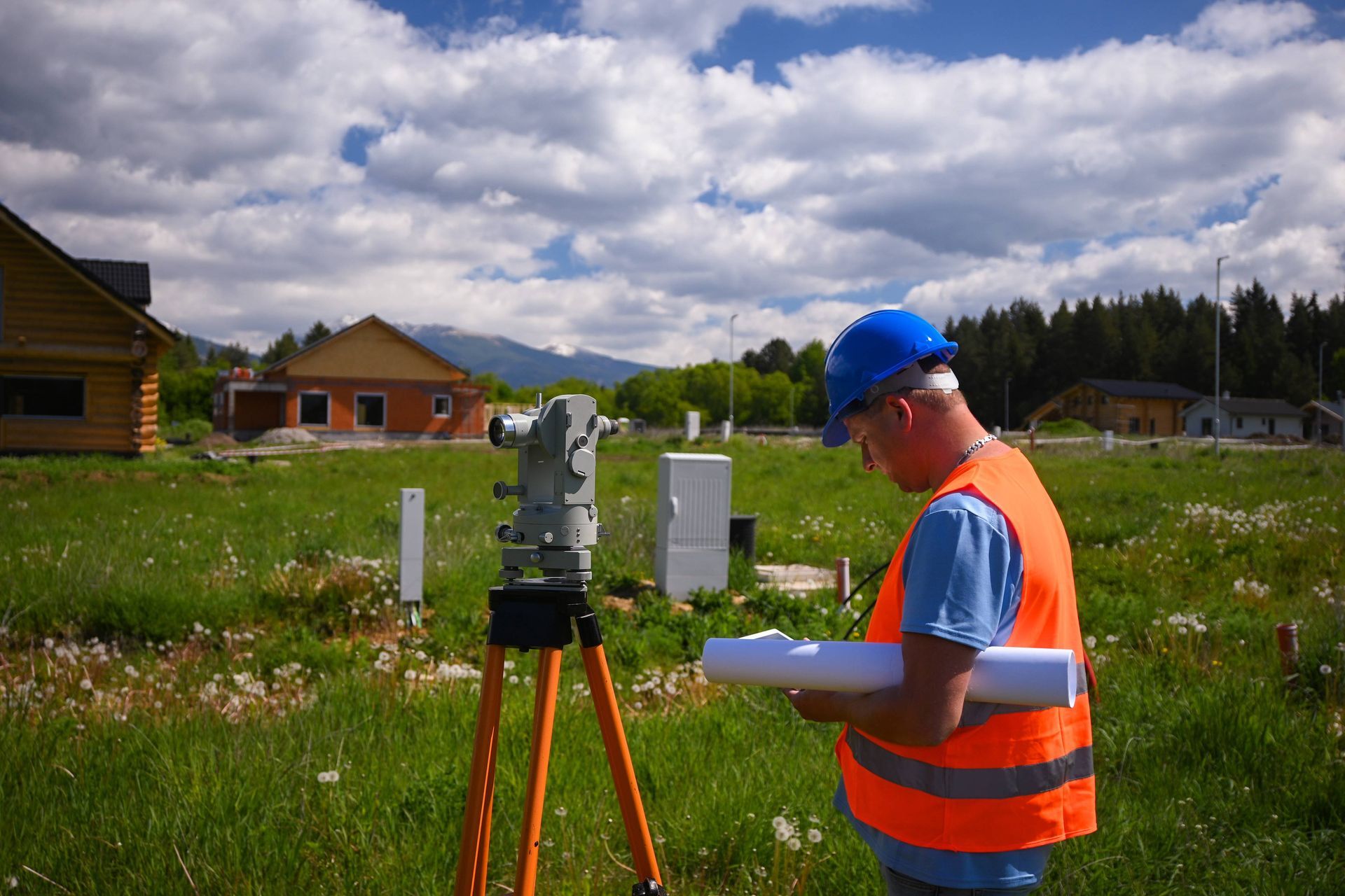 Surveyor in orange vest and blue hard hat checks a tripod instrument in a grassy field near houses Surveyor in orange vest and blue hard hat checks a tripod instrument in a grassy field near houses
