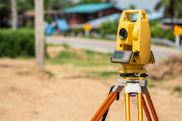 Yellow surveying instrument on a tripod outdoors near a dirt road.