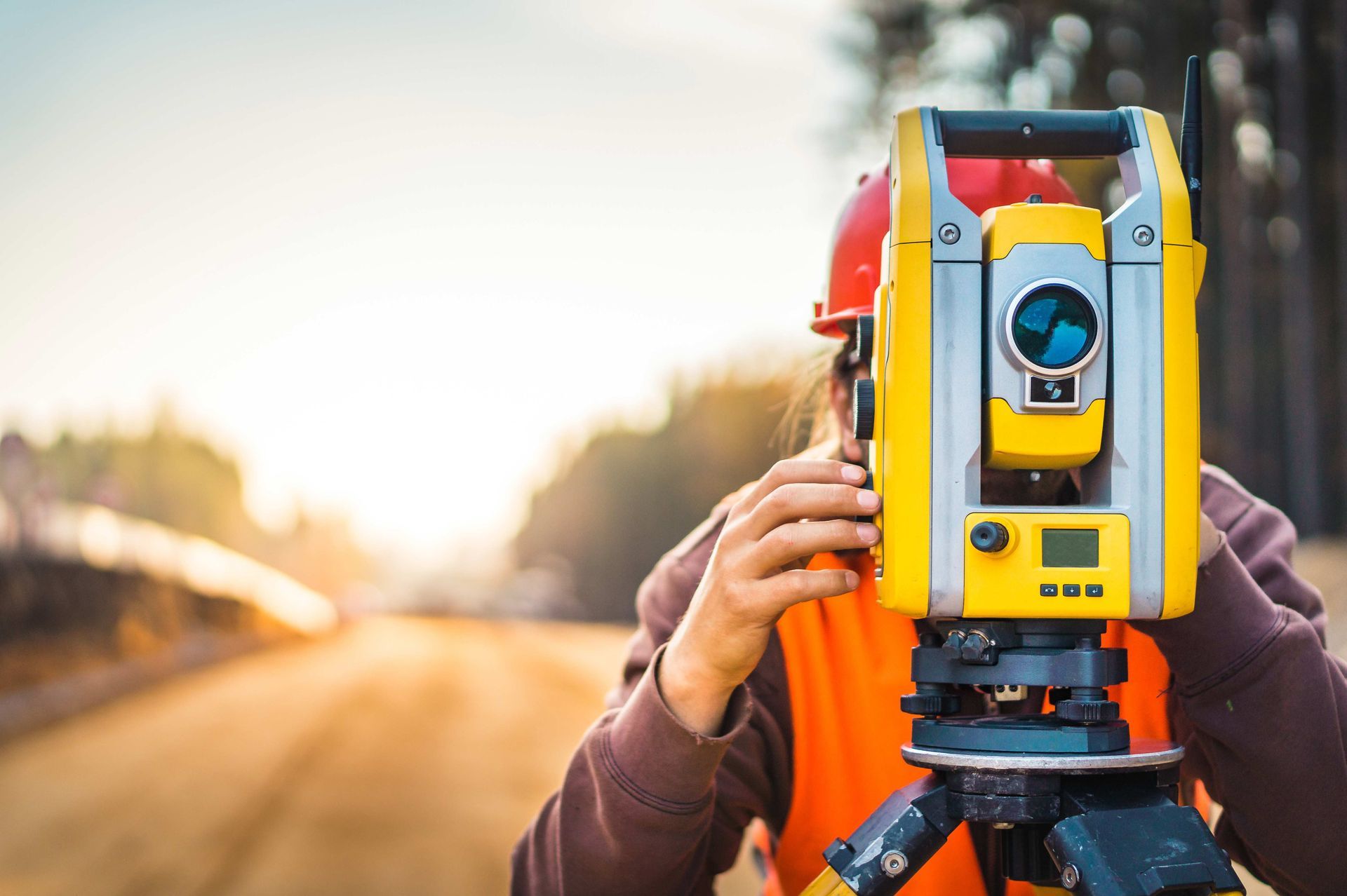 Person using a yellow surveyor’s tool on a tripod outdoors at sunset Person using a yellow surveyor’s tool on a tripod outdoors at sunset