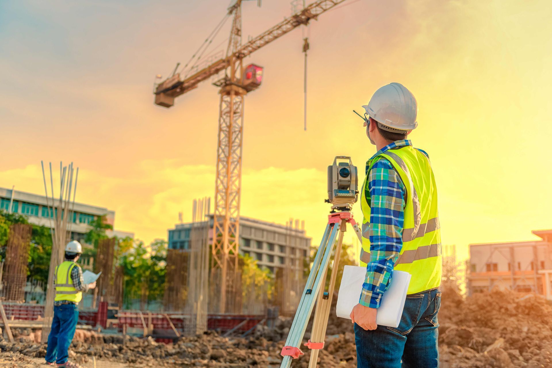 Construction workers surveying a site at sunset with a crane and tripod-mounted measuring equipment
