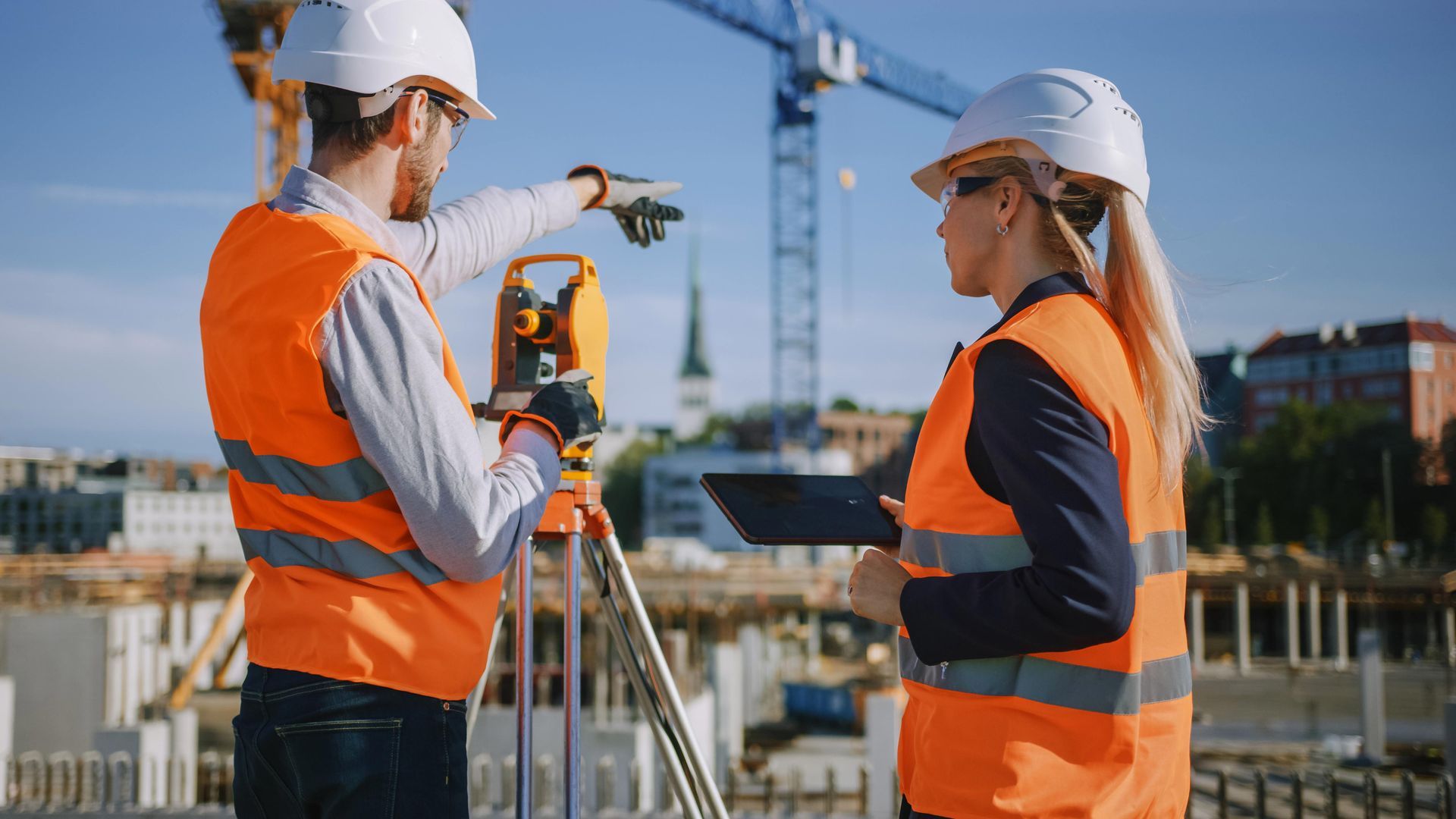 Two construction workers in orange safety vests survey a site with surveying equipment near a building foundation.