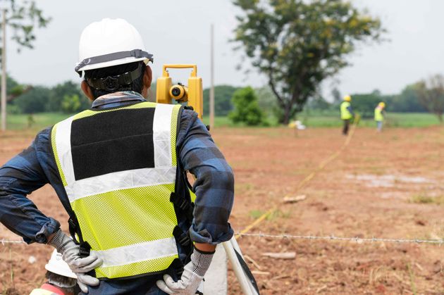 Surveyor in white hard hat and safety vest using equipment on a construction site, with fields in background