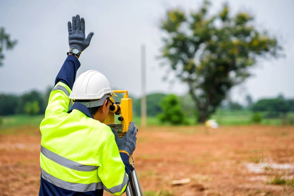 Surveyor in hard hat and reflective jacket using a total station in a field, one hand raised.