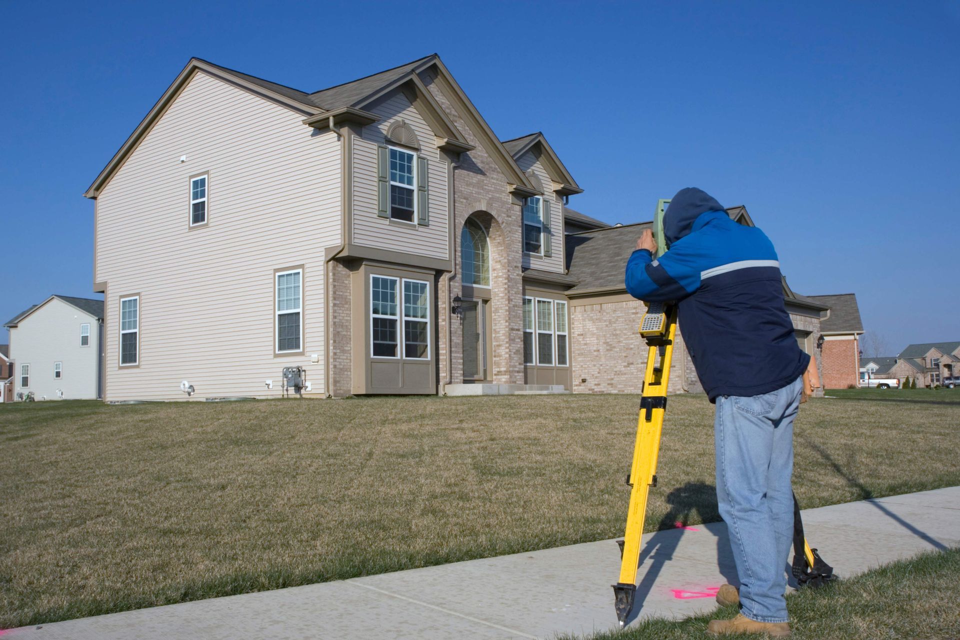 Worker using a yellow survey tool in front of a beige suburban house under construction Worker using a yellow survey tool in front of a beige suburban house under construction