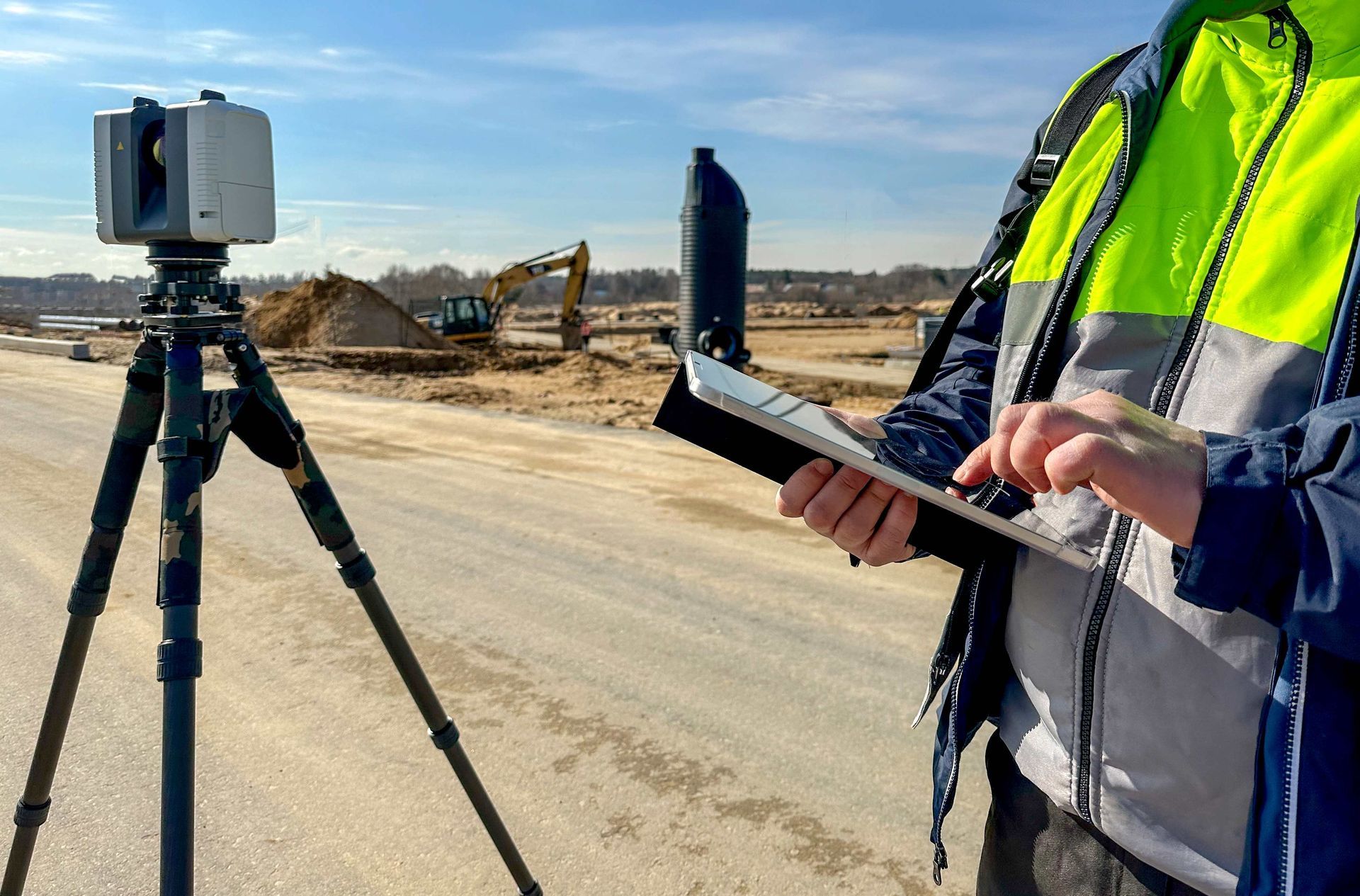 Surveying equipment on a tripod at a construction site, with a worker holding a tablet nearby.