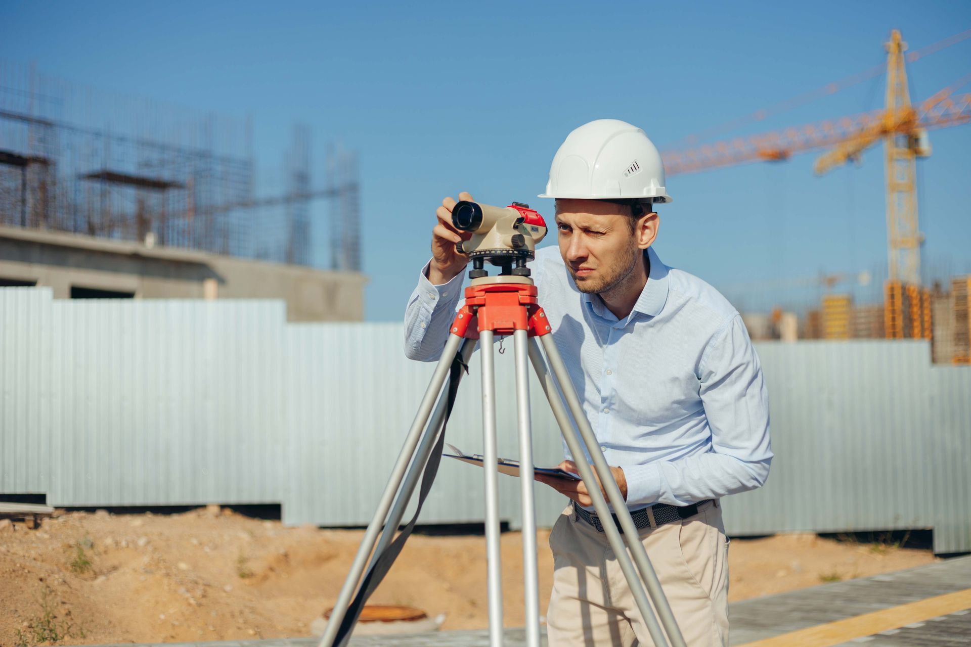 Surveyor using a tripod-mounted instrument on a construction site, with cranes and building materials in the background