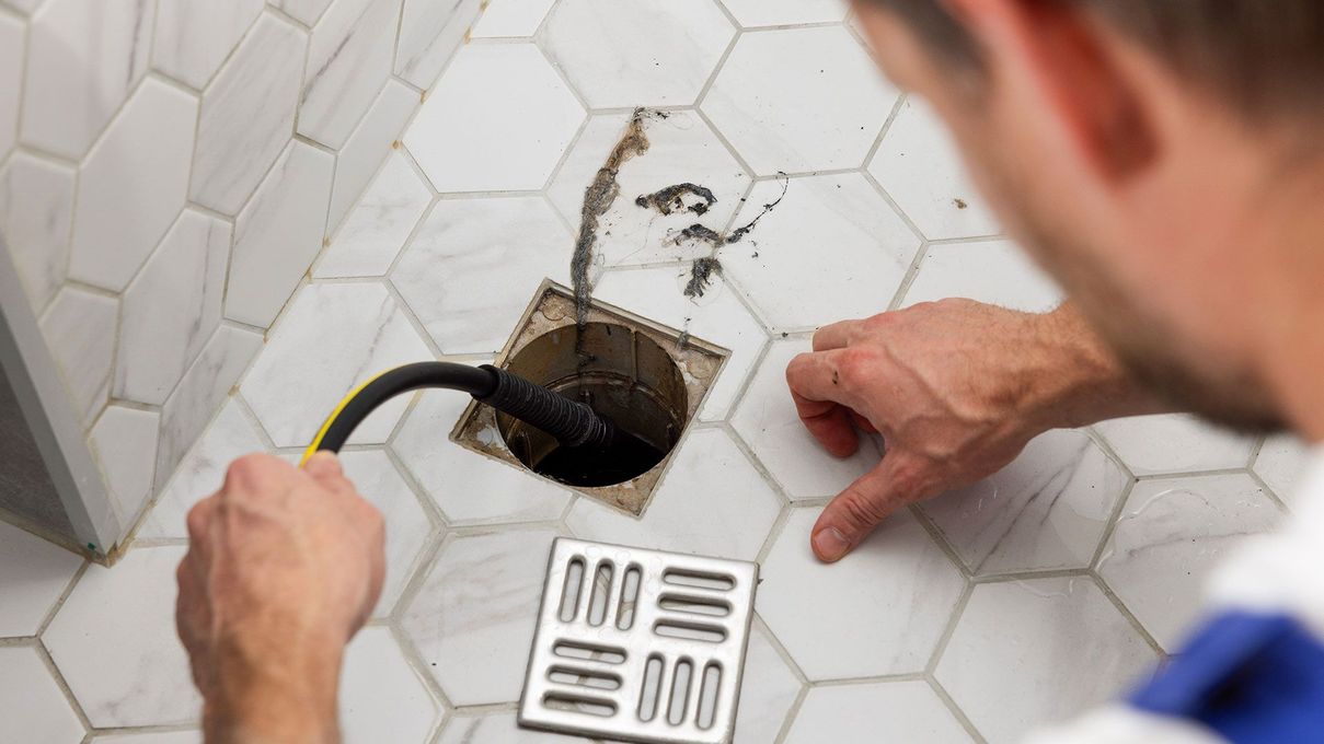 Plumber unclogging a floor drain in white tiled bathroom with a hose and drain cover